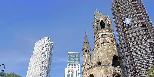 Kurfürstendamm  Im Hintergrund sind moderne Gebäudetürme zu sehen die sich in den Himmel erstrecken. Im vorderen Teil des Bildes ist die durch den 2. Weltkrieg zerstörte Kaiser-Wilhelm-Gedächtniskirche zu sehen.