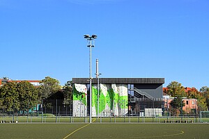 Ein modernes Kletterzentrum mit einer grünen, auffälligen Kletterwand und einem großen Sportplatz davor. Der blaue Himmel hebt die klare Struktur des Gebäudes hervor.