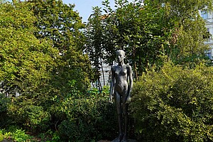 Eine schlanke Bronzestatue einer Frau steht zwischen dicht bewachsenen Büschen und Bäumen im Helmholtzplatz. Die Statue zeigt eine stehende Frau mit ernstem Blick und schlichten Zügen. Rundherum wuchert grünes Laub, und ein Gebäude mit weißen Fassaden schimmert im Hintergrund durch die Blätter.