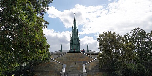Monument im Viktoriapark Ein großes Denkmal mit grünem, gotischem Spitzdach erhebt sich auf einem breiten Sockel, umgeben von Bäumen im Viktoriapark in Berlin. Mehrere Stufen führen hinauf, und einige Besucher sitzen auf der Plattform unter dem Denkmal.