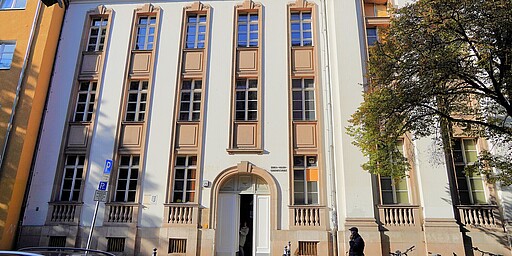 Fassade der Erik-Mann-Grundschule in Berlin Eine helle Fassade der Erik-Mann-Grundschule in Berlin. Das Gebäude ist mehrstöckig mit hohen, schmalen Fenstern und dekorativen Elementen um die Fenster herum. Links ein gelbes Nebengebäude, rechts ein Baum mit Herbstlaub. Auf der Straße sind einige Fahrräder und eine Person zu sehen.