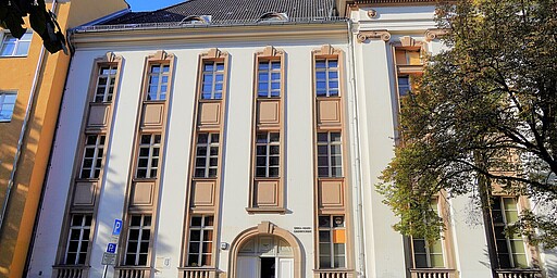 Fassade der Erik-Mann-Grundschule in Berlin Eine helle Fassade der Erik-Mann-Grundschule in Berlin. Das Gebäude ist mehrstöckig mit hohen, schmalen Fenstern und dekorativen Elementen um die Fenster herum. Links ein gelbes Nebengebäude, rechts ein Baum mit Herbstlaub. Auf der Straße sind einige Fahrräder und eine Person zu sehen.
