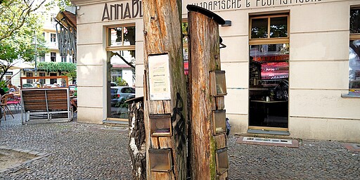 Bücherwald vor dem Café Anna Blume  Das Bild zeigt den "Bücherwald" vor dem Café Anna Blume im Prenzlauer Berg, Berlin. Der Bücherwald besteht aus mehreren aufrecht stehenden Baumstämmen, in die kleine Fächer eingelassen sind, die Bücher enthalten. Diese Art von öffentlicher Büchertauschbörse ermöglicht es Passanten, kostenlos Bücher mitzunehmen oder eigene hineinzustellen. Im Hintergrund sieht man die Fassade des Cafés Anna Blume, das nach Kurt Schwitters' berühmtem Gedicht benannt ist.