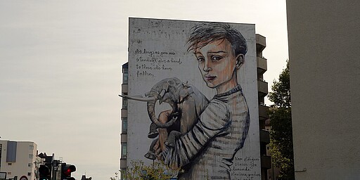 Wandgemälde über Mitgefühl und Gemeinschaft Ein Wandgemälde zeigt einen jungen Menschen, der einen Elefanten umarmt. Die Inschrift fordert dazu auf, anderen zu helfen, die gefallen sind, solange man selbst steht.