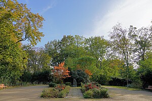 Eine weitläufige Parkfläche mit einem zentralen Blumenbeet und buntem Herbstlaub. Im Hintergrund stehen hohe Bäume, die in warmen Gelb-, Rot- und Grüntönen leuchten. Zwei Holzbänke stehen am Rand des Weges, der sich durch den Park schlängelt.