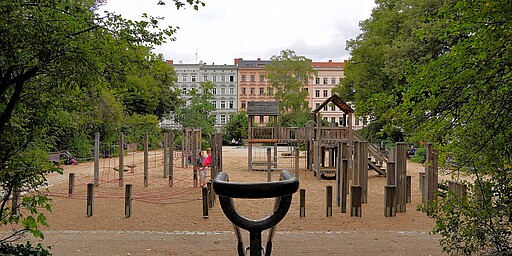 Holzspielplatz am Chamissoplatz Ein Spielplatz am Chamissoplatz in Kreuzberg mit Fokus auf die Holzspielgeräte. Im Vordergrund sind Holzpfosten und ein Klettergerüst mit Netzen zu sehen. Dahinter ragen pastellfarbene Altbauten in die Höhe, die dem Platz ein klassisches Berliner Flair verleihen. Kinder und Eltern genießen die Zeit im Sandbereich.