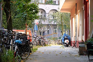 Ein ruhiger Gehweg in der Göhrener Straße im Prenzlauer Berg, Berlin. Links stehen viele geparkte Fahrräder, ein Roller ist am rechten Rand geparkt, und im Hintergrund sind moderne, gebogene Spielplatzstrukturen zu sehen, bekannt als das Göhrener Ei. Die Bäume und das helle Tageslicht verleihen der Szene ein entspanntes Stadtgefühl.