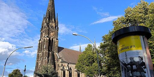 Gotische Fassade der Kirche am Südstern Eine imposante Kirche mit gotischer Architektur und einem hohen spitzen Turm. Der Himmel ist strahlend blau, und im Vordergrund sieht man eine Werbesäule mit einem gelben Plakat.