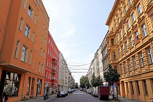 Eine schmale Straße mit bunten Wohnhäusern auf beiden Seiten, darunter orange, rote und gelbe Fassaden. Einige Gebäude haben aufwendige Verzierungen. Über der Straße hängen kleine Girlanden, und am Rand parken Autos.