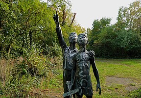 Eine Skulptur von zwei Menschen auf dem Syringenplatz. Die Figuren, schwarz bemalt und mit verschiedenen Mustern versehen, stehen eng beieinander. Eine der Figuren hebt den Arm und zeigt in die Ferne, während die andere mit einem Arm um die Schulter des ersten steht. Die Szene ist umgeben von dichter Vegetation mit hohen Bäumen und Büschen, die einen ruhigen, naturverbundenen Eindruck vermitteln.