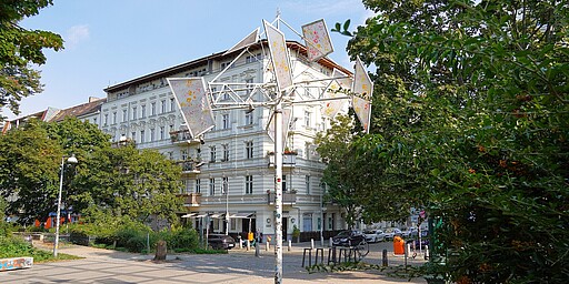 Kunstinstallation auf dem Helmholtzplatz  Eine kunstvolle Installation auf dem Helmholtzplatz, bestehend aus metallischen Strukturen, die wie bunte Flaggen im Wind wirken. Die Skulptur steht vor einem alten, weißen Gebäude mit verzierter Fassade. Der Platz ist von Bäumen umgeben, und im Vordergrund sind Büsche und Blumen zu sehen, die das Grün der Umgebung ergänzen.