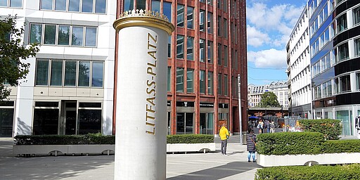 Litfaßsäule am Litfaßplatz in Berlin Eine weiße Litfaßsäule mit goldener Krone und der Aufschrift "Litfaß-Platz" steht inmitten eines modernen Platzes mit Bürogebäuden im Hintergrund. Einige Passanten flanieren und beleben die Szene.