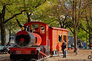 Ein leuchtend roter Holzzug, liebevoll gestaltet mit der Aufschrift Zauberschul-Express, steht auf einem Spielplatz unter Bäumen mit herbstlich gefärbten Blättern. Kinder spielen in und um den Zug.