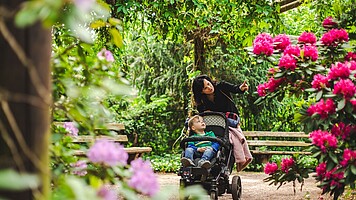 Eine junge Frau mit Kinderwagen zeigt ihrem Kind die bunten Blüten der Rhododendronpflanze