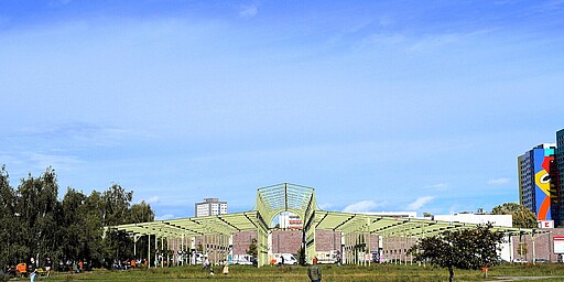 Die alte Hammelauktionshalle im Blaneksteinpark Weitläufiger Blick auf eine grüne Wiese mit vereinzelten Spaziergängern, im Hintergrund eine große, offene Eisenkonstruktion, die früher als Hammelauktionshalle genutzt wurde. Links und rechts sind Bäume zu sehen, der Himmel ist klar und blau.