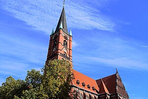 Die beeindruckende rote Backsteinkirche mit einem hohen Kirchturm, der in den blauen Himmel ragt. Der Turm ist mit grünen Spitzen und Ornamenten geschmückt. Im Vordergrund befindet sich eine Baumreihe, deren Blätter teilweise das Gebäude verdecken.