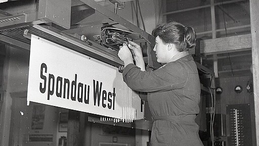 Eine DB‑Mitarbeiterin arbeitet an einem Elektronikmodul der Bahnhofsanzeige in Spandau West – ein Symbol für den Einstieg von Frauen in technische Berufe bei der Bahn.