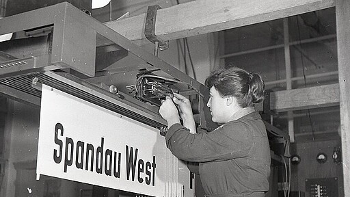 Eine DB‑Mitarbeiterin arbeitet an einem Elektronikmodul der Bahnhofsanzeige in Spandau West – ein Symbol für den Einstieg von Frauen in technische Berufe bei der Bahn.
