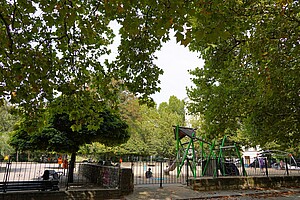 Ein moderner Spielplatz mit grünen Klettergerüsten, einer Rutsche und Sandboden. Im Vordergrund hängt eine grüne Baumkrone. Eltern und Kinder sind am Spielen.