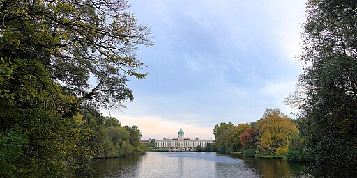 Blick auf das Schloss Charlottenburg über den Schlossgarten-Teich  Eine ruhige Szene im Schlossgarten Charlottenburg: Im Vordergrund ein stiller Teich, umgeben von Bäumen mit herbstlichem Laub. Im Hintergrund thront das imposante Schloss Charlottenburg mit seiner markanten grünen Kuppel, eingebettet in die Natur.