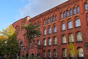 Ein imposantes, historisches Gebäude aus rotem Backstein mit großen Fenstern und detailreichen Verzierungen. Bäume mit Herbstlaub stehen davor, und der Himmel ist strahlend blau.