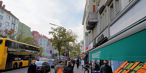 Lebendiges Straßenleben an der Turmstraße Das Bild zeigt die Turmstraße in Berlin. Links im Vordergrund steht ein auffälliger gelber Doppeldeckerbus mit der Aufschrift "Sanssouci.de". Rechts sind Marktstände und Geschäfte, darunter ein Obstladen mit leuchtenden Farben. Menschen schlendern die Straße entlang, während im Hintergrund moderne und klassische Wohnhäuser zu sehen sind.