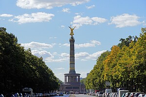 Die Berliner Siegessäule erhebt sich majestätisch in die Höhe, mit ihrer goldenen Statue, die in der Sonne glänzt. Sie wird von einer Allee mit Bäumen und parkenden Autos eingerahmt.