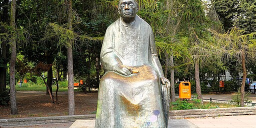 Skulptur von Käthe Kollwitz auf dem Kollwitzplatz  Eine bronzene Skulptur von Käthe Kollwitz sitzt auf einem Sockel im Kollwitzplatz. Sie wirkt nachdenklich, hält ein kleines Buch oder Objekt in der Hand und ist von Bäumen und Grün umgeben. Die Skulptur ist leicht verwittert, was ihr eine besondere Atmosphäre verleiht.