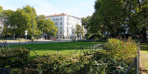 Blick auf den Savignyplatz in Berlin  Ein ruhiger Blick auf den Savignyplatz in Berlin. Im Vordergrund befindet sich üppige Vegetation, hinter einem kleinen Zaun sieht man saftig grünen Rasen. Im Hintergrund erheben sich Altbauten mit weißen Fassaden, umgeben von dichten Bäumen.