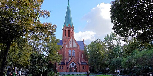 Herbstliche Ruhe vor der St.-Ludwig-Kirche  Auf dem Bild ist die St.-Ludwig-Kirche in Berlin zu sehen, eine große neugotische Backsteinkirche mit einem hohen grünen Turm, der spitz in den Himmel ragt. Der Vorplatz der Kirche ist von Bäumen gesäumt, die herbstlich verfärbt sind, und es gibt einen Brunnen. Ein paar Menschen spazieren entspannt entlang, die Atmosphäre ist ruhig und friedlich.