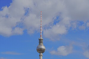 Das Bild zeigt den Berliner Fernsehturm, das höchste Gebäude der Stadt, vor einem hellblauen Himmel mit weißen Wolken. Die Kugel mit ihrer glänzenden Oberfläche und die rot-weiß gestreifte Antenne ragen majestätisch in die Höhe.