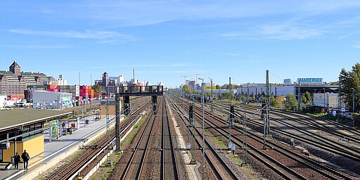 Weitläufige Gleisanlage am Westhafen Ein Blick über die Gleisanlagen der S-Bahn-Station Westhafen in Berlin. Die Schienen verlaufen in parallelen Linien in die Ferne. Im Hintergrund sind moderne und historische Lagerhäuser sowie bunte Containerstapel zu sehen. Der Himmel ist klar und blau, ein sonniger Tag.