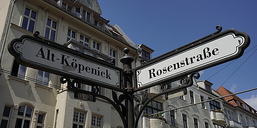  Ein dekoratives Straßenschild im historischen Stil zeigt die Straßennamen „Alt-Köpenick“ und „Rosenstraße“. Im Hintergrund sind helle Altbauten mit hohen Fenstern und teils rot gedeckten Dächern zu sehen, die das typische Bild der Altstadt Köpenicks widerspiegeln. 