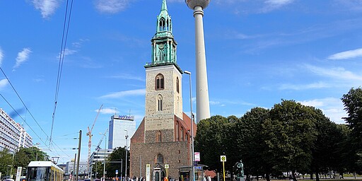 St. Marienkirche und Berliner Fernsehturm an der Karl-Liebknecht-Straße Eine Ansicht der St. Marienkirche in Berlin mit ihrem markanten Kirchturm aus hellem Stein und grüner Spitze, daneben erhebt sich der Fernsehturm, der sich in den blauen Himmel erstreckt. Im Vordergrund fahren Straßenbahnen und Autos an der Straße vorbei, während Menschen den Gehweg entlang laufen.