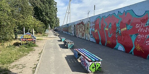 Graffiti-Kunst im Mauerpark  Ein Spazierweg entlang der ehemaligen Berliner Mauer im Mauerpark. Die Mauer ist mit bunten Graffiti-Kunstwerken bedeckt, die sich über ihre gesamte Länge erstrecken. Auf der linken Seite stehen Bäume und eine Sitzbank, die ebenfalls mit Graffiti bemalt ist. Die Atmosphäre ist kreativ und lebendig.