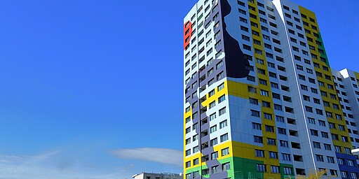  Ein hohes Gebäude mit einer farbenfrohen Fassade. Auf der rechten Seite sieht man eine große schwarze Form, die an ein abstraktes Gesicht erinnert. Die unteren Etagen sind gelb, grün und violett, während der Himmel im Hintergrund tiefblau ist. 