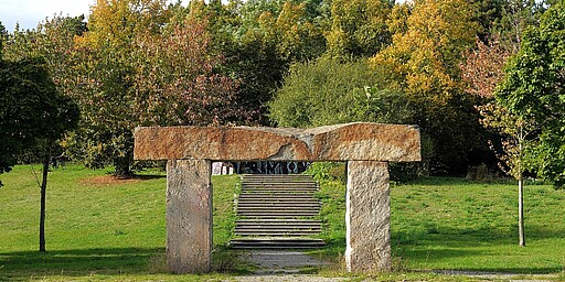  Im Vordergrund ein massives Steintor, das aus zwei aufrecht stehenden Steinblöcken und einem darauf liegenden Querbalken besteht. Dahinter führen mehrere Stufen hinauf, eingerahmt von Bäumen in herbstlichen Farben. Der Himmel ist leicht bewölkt. 