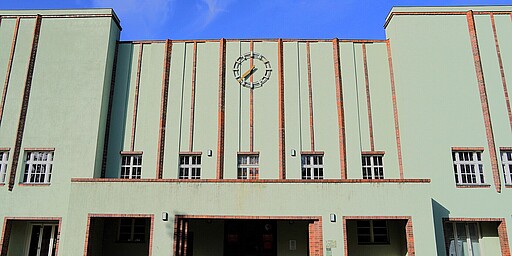 Klassische Stadionuhr Die Fassade des Poststadions, hellgrün gestrichen mit orangefarbenen Backstein-Details. Über dem Eingang thront eine große, runde Uhr. Der Himmel ist strahlend blau.