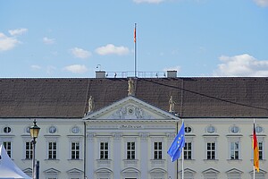 Das Bild zeigt Schloss Bellevue, die offizielle Residenz des deutschen Bundespräsidenten. Die elegante weiße Fassade ist mit Statuen und Ornamenten geschmückt. Über dem Dach weht die Flagge des Bundespräsidenten, daneben die Europa- und die Deutschlandflagge.