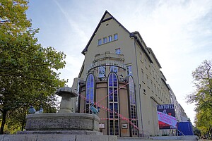 Ein historisches Gebäude mit großen Fenstern und einem markanten Eingang. Herbstliche Bäume säumen den Platz, ein großes Schild weist auf eine Veranstaltung hin.