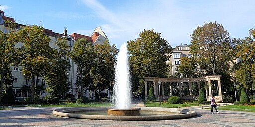  Ein sonniger Tag am Viktoria-Luise-Platz in Berlin. Ein großer, runder Brunnen mit sprudelndem Wasser steht im Mittelpunkt des Platzes. Um den Brunnen herum gibt es gepflegte Grünflächen und Bäume, während Wohnhäuser den Hintergrund schmücken. 
