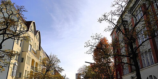 Herbstliche Häuser im Bayerischen Viertel  n einer herbstlichen Szenerie mit klarblauem Himmel stehen rechts und links zwei Altbau-Häuser. Die Bäume an der Straße tragen teilweise gelbes Laub, das die herbstliche Stimmung unterstreicht. Die Fassaden der Gebäude sind in hellen Farben gehalten, und die Bäume werfen Schatten auf die Gehwege. Im Hintergrund ist das Rathaus Schöneberg zu erkennen.