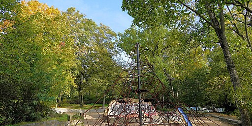 Ein Spielplatz im Park, umgeben von dichten, herbstlich gefärbten Bäumen. Im Mittelpunkt steht ein Klettergerüst in Form einer Pyramide mit roten Seilen, an dem Kinder klettern können. Der Boden ist mit Sand bedeckt.