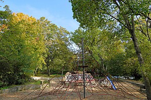 Ein Spielplatz im Park, umgeben von dichten, herbstlich gefärbten Bäumen. Im Mittelpunkt steht ein Klettergerüst in Form einer Pyramide mit roten Seilen, an dem Kinder klettern können. Der Boden ist mit Sand bedeckt.