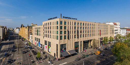 Modernes Shopping im Schultheiss Quartier Das Schultheiss Quartier aus der Vogelperspektive mit seinem modernen Einkaufszentrum aus beigefarbenen Steinen. Die große Glasfassade mit Geschäften und Cafés ist von Passanten und Fahrrädern belebt. Die Straßen davor sind gut befahren.