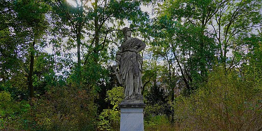 Borussia-Statue im Preußenpark  Eine steinerne Statue der Göttin Borussia, die in einer majestätischen Pose im Preußenpark steht. Sie trägt eine Rüstung und einen Helm, der in ihrer linken Hand ruht, während sie in der rechten Hand einen Lorbeerkranz hält. Im Hintergrund umgeben dichte grüne Bäume die Statue, durch die das Sonnenlicht strahlt.