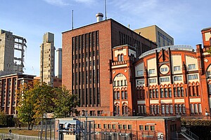 Ein großes industrielles Gebäude aus rotem Backstein und Beton, daneben eine im Abriss befindliche Struktur. Bäume in herbstlichen Farben säumen die Umgebung.