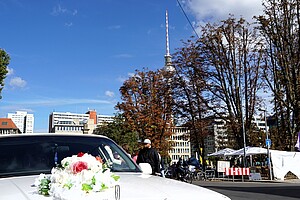 Ein geschmücktes weißes Auto mit einem Blumengesteck auf der Motorhaube steht im Vordergrund. Im Hintergrund ragen Bäume und der Berliner Fernsehturm in den blauen Himmel. Menschen gehen vorbei, und rechts sind weiße Zelte zu erkennen, vermutlich Teil einer Veranstaltung oder eines Marktes.