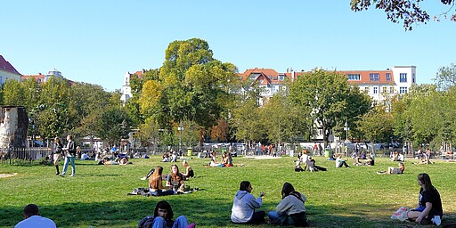 Boxhagener Platz Sicht auf den Boxhagener Platz von Osten. Menschen sitzen auf einer grünen Wiese in einem belebten Park. Viele genießen das sonnige Wetter, während sie im Gras entspannen oder sich unterhalten. Im Hintergrund sind Bäume sowie helle Wohnhäuser mit roten Dächern zu sehen.