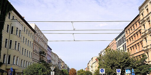 Oderberger Straße mit Blick auf Berliner Fassaden  Eine malerische Straße in Berlin, umrahmt von Altbauten in hellen Farben. Im Vordergrund gibt es viele Pflanzen und gemütliche Straßencafés. Die warme Atmosphäre lädt zum Verweilen ein.